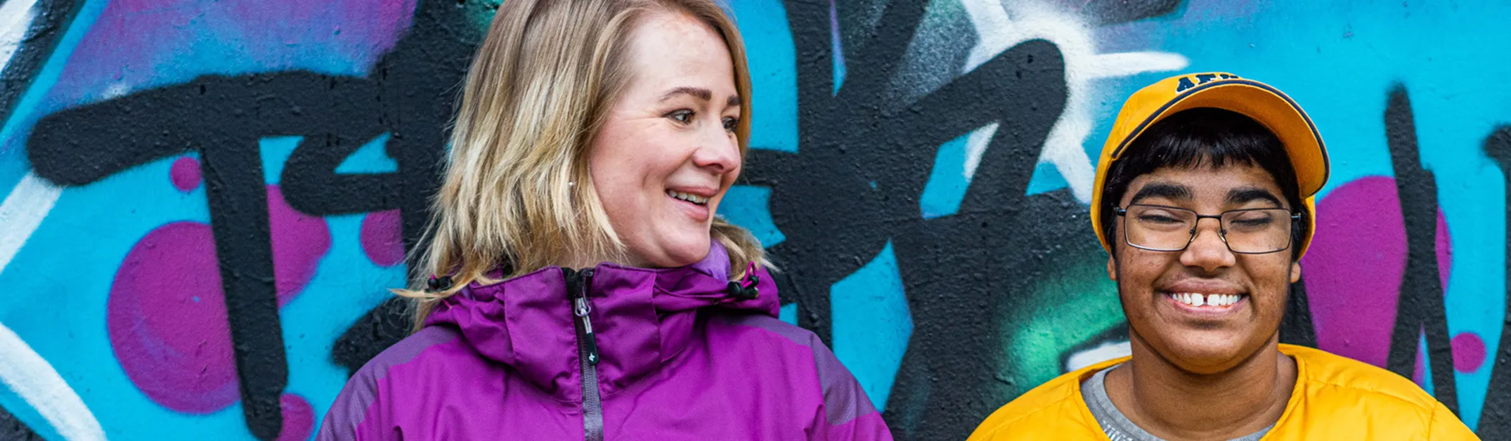 Personlig assistans hos JAG. På bilden tittar den personliga assistenten mot assistansanvändaren, bakgrunden är en färgglad grafittivägg utomhus.