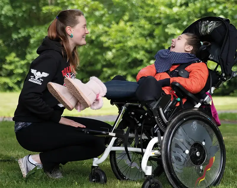 En flicka i rullstol på en gräsmatta. Hon som tillhör personkrets 1 av  tre personkretsar Hennes assistent sitter på huk framför henne.