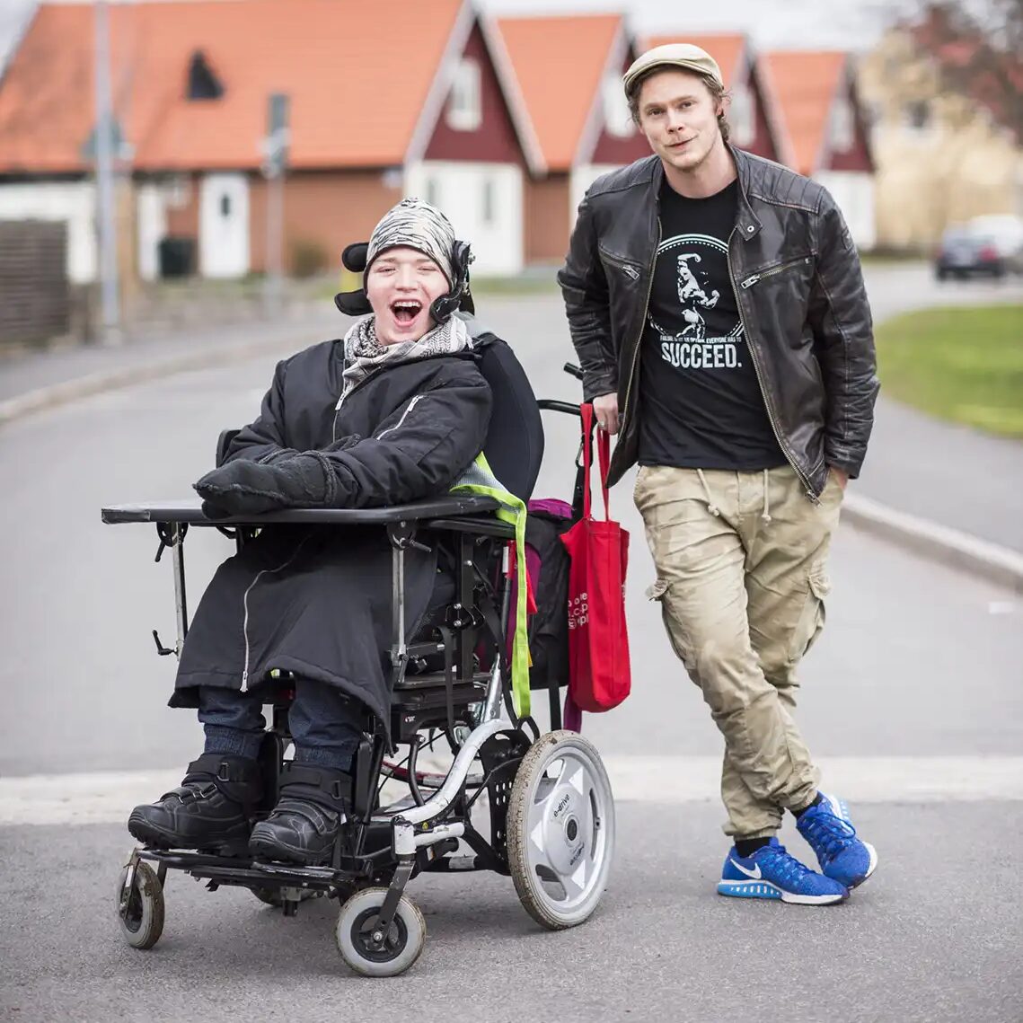 En skrattande man i rullstol med sin assistent stående bredvid på en asfalterad villagata.