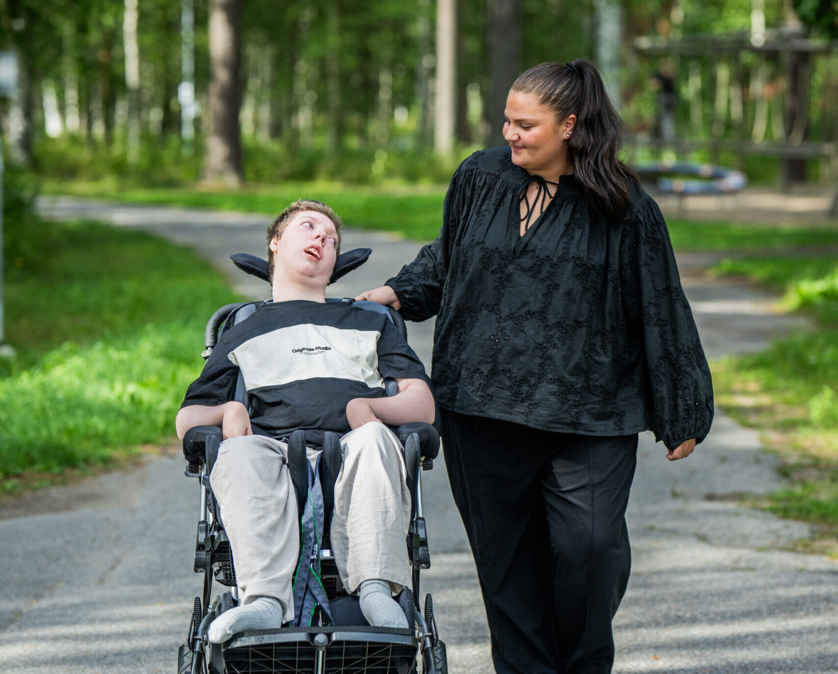 Assistansanvändare i rullstol och hens assistent promenerar bland grönska. 