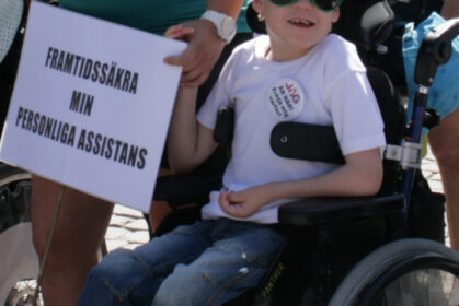 Foto av ett barn i rullstol med en skylt där det står "Framtidssäkra assistansen"