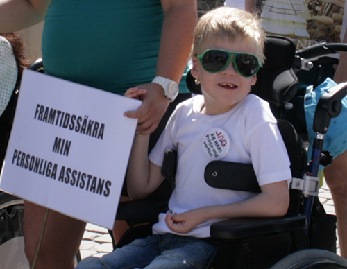 Litet barn med solglasögon sitter i en rullstol. En skylt med texten Framtidssäkra min assistans.