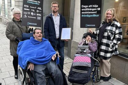 Fem personer i ytterkläder framför en pampig entré. Två av personerna är rullstolsburna. En av personerna håller i en pappkartong.