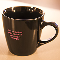 Svart kaffemugg med rosa text: "Även om jag inte säger något kan jag ha något att säga"