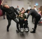 En kvinna i rullstol omgiven av två dansande personer. I bakgrunden fler personer som dansar.