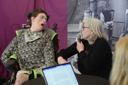 En ung man i rullstol och en kvinna som sitter bredvid. Hon pratar i en mikrofon.