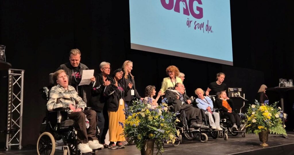 Många glada och finklädda människor på en scen, några i rullstol. 