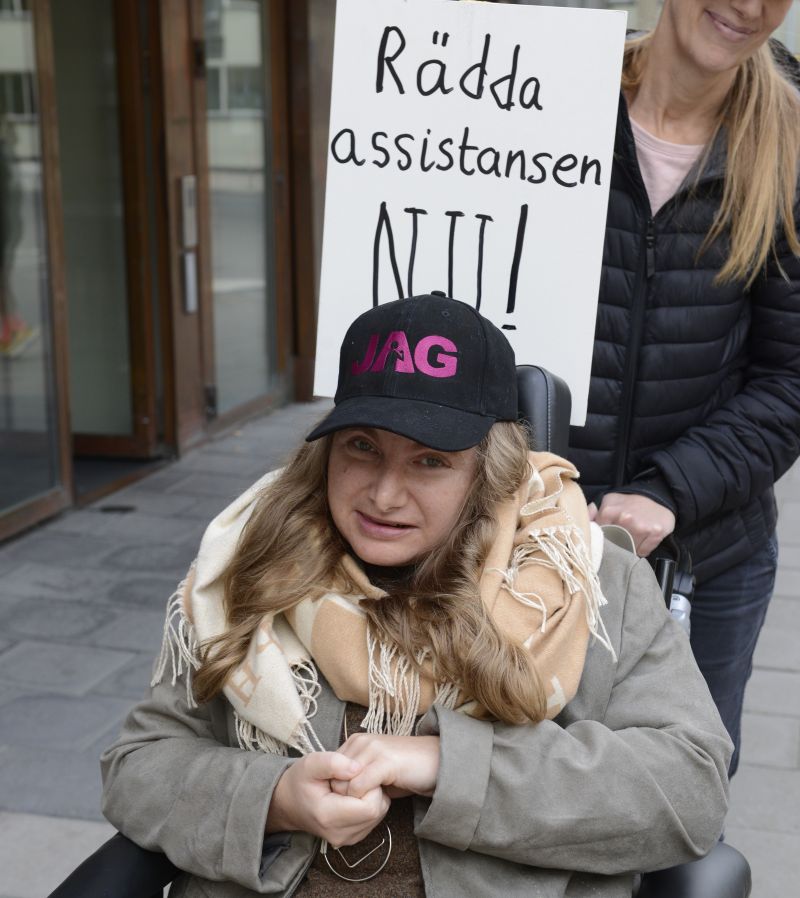 Rullstolsburen kvinna med keps med JAGs logga med demonstrationsskylt Rädda assistansen NU!