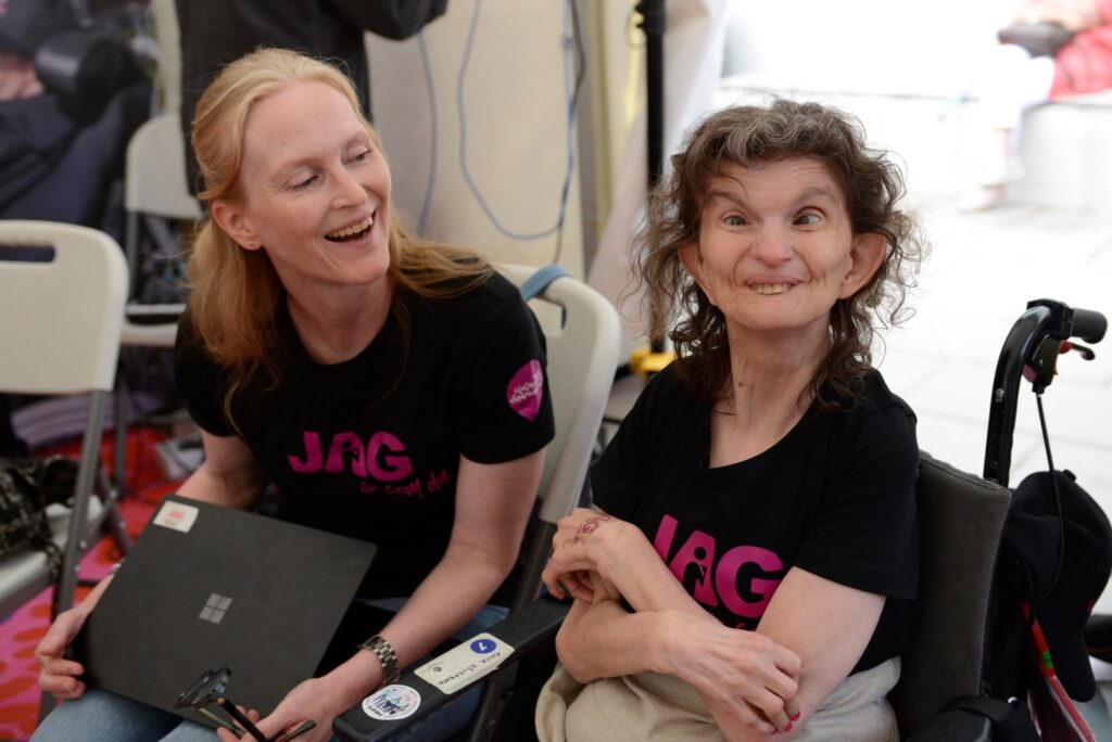 Två leende kvinnor i svart t-shirtar med JAGs rosa logga.