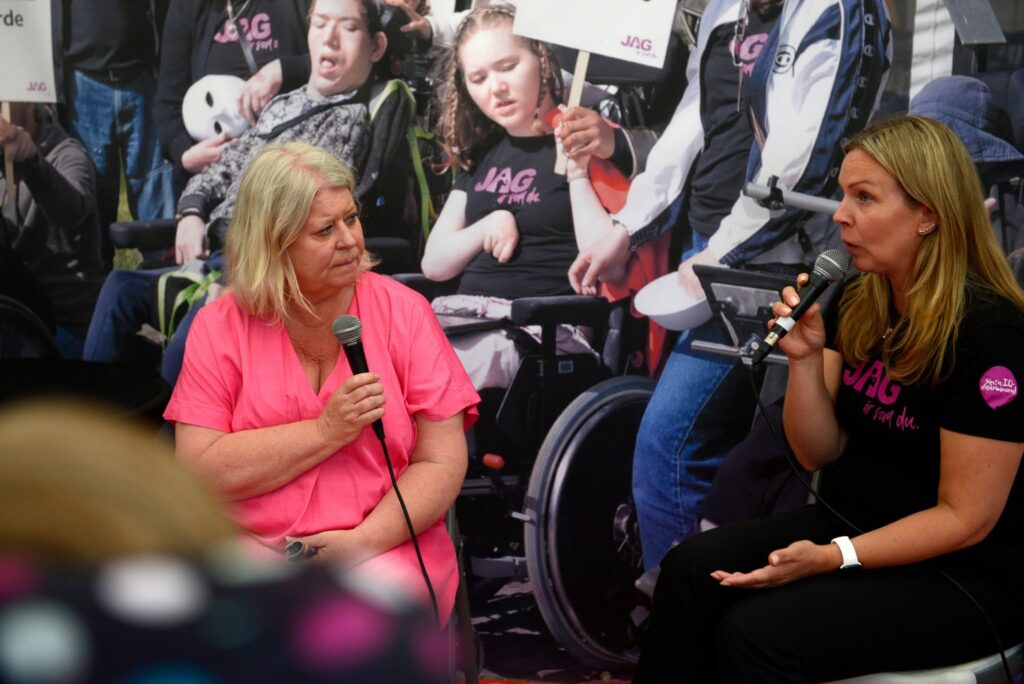 Socialministern i rosa klänning intervjuas framför en fondvägg med ett foto av JAG-medlemmar med demonstrationsskyltar.